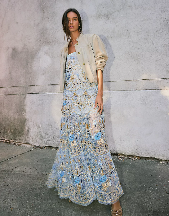A person stands on a sidewalk against a textured, light gray wall, wearing a light tan jacket over a blue and beige boho maxi dress with floral designs—perfect for weekend getaways. They have one hand on their hip and are looking at the camera.