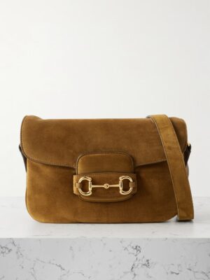 A brown suede shoulder bag inspired by the Gucci Horsebit 1955 Soft Small Leather Tote Bag, with a flap closure, gold hardware detail on the front, and a matching suede strap, displayed on a white marble surface against a light background.