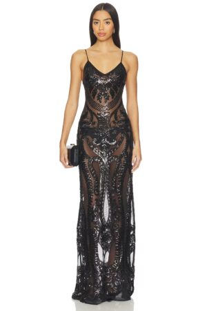 A woman stands facing forward wearing a long, black Norma Kamali Low Back Fara Slip Fishtail Gown with intricate sequin patterns. The dress has thin straps and a fitted silhouette. She holds a small black clutch and has straight, loose hair.