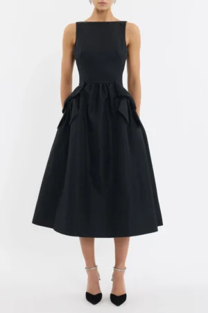 A woman wears the Rebecca Vallance Narcissa Mini Dress, a sleeveless black piece with a fitted bodice, flared skirt, and bows at the hips. She pairs it with black pointed-toe heels with ankle straps against a plain white background.