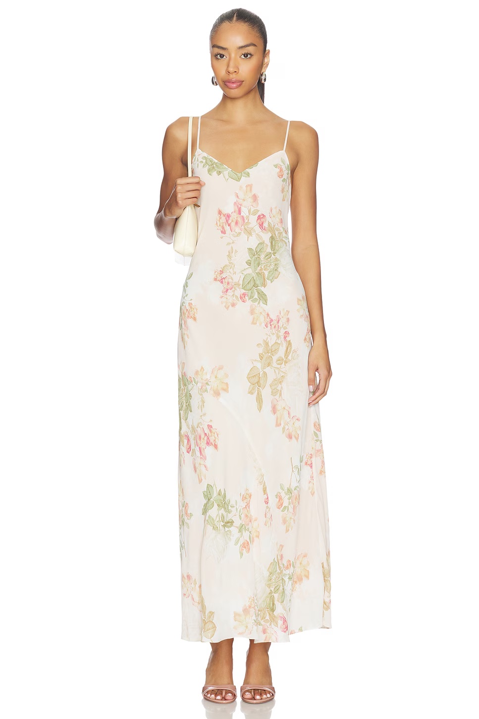 A woman stands facing forward in a long, sleeveless spring floral dress with a light pattern. She carries a small white purse, wears sandals, and has her hair tied back with a neutral expression.