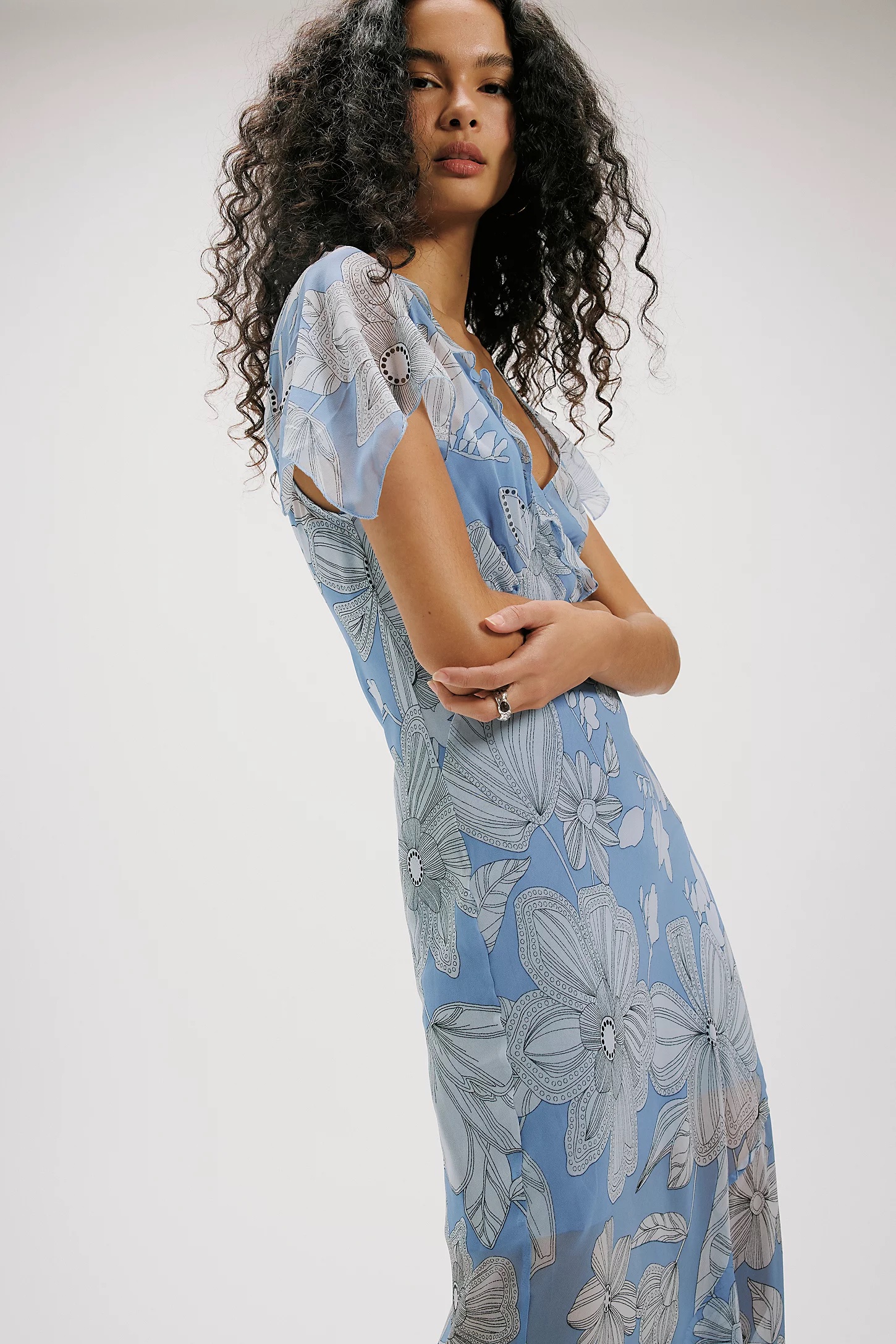 A person with long, curly hair wears a light blue, short-sleeved spring floral dress with large white patterns, standing against a plain, light-colored background.
