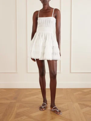 A person stands indoors on a wooden floor, wearing an Alaïa Off-Shoulder Sculpting Line Flared Wool Mini Dress with thin straps and horizontal stripe details, paired with white flat sandals. The background features cream-colored paneled walls.