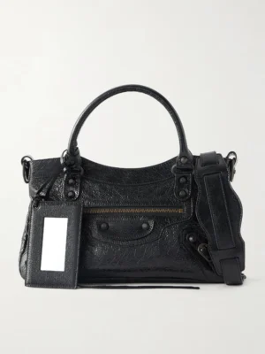 A black textured leather City Mini Bag with two top handles, a front zipper pocket, silver-tone hardware, an attached rectangular tag, and a detachable shoulder strap. Charms add a playful accent to this chic bag displayed against a plain white background.