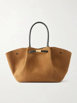 A large tan suede tote bag with black leather handles and a gold-tone clasp at the top center. The Demellier The New York Leather-trimmed Suede Tote features a soft, structured shape and minimal branding near the base.