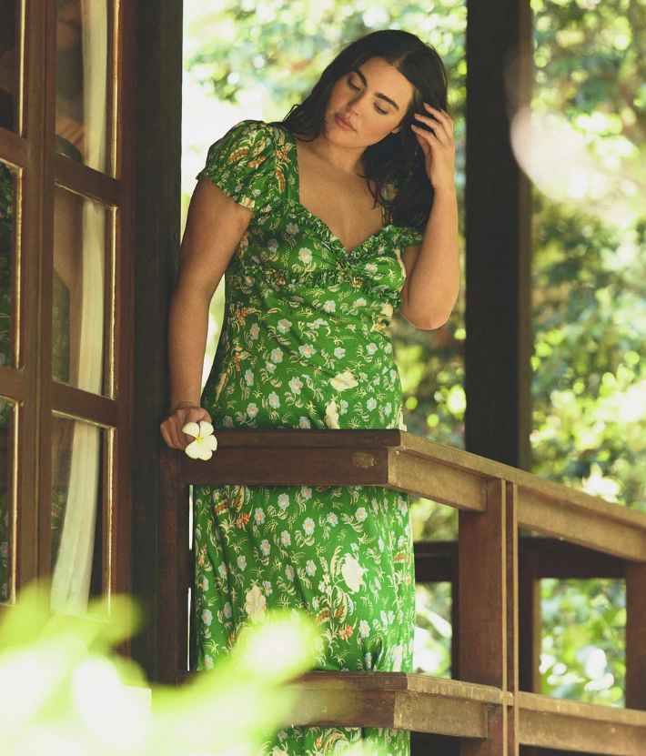 A woman in a green floral French inspired summer dress stands on a wooden porch, holding a white flower in one hand and touching her hair with the other. The background shows trees and greenery.