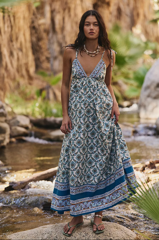 A woman stands outdoors by a stream, wearing a long, patterned sundress with blue and white floral summer dress designs. She has long hair, wears sandals, layered necklaces, and is surrounded by green plants and rocks.