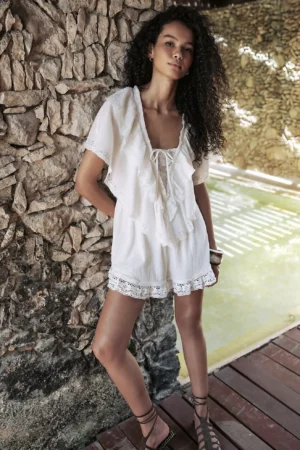 A person with long curly hair stands against a textured stone wall, wearing a white, short-sleeve romper with lace trim and a tie front, reminiscent of the Free People Audra Midi Dress. Wooden decking and a sunlit pool are visible in the background.