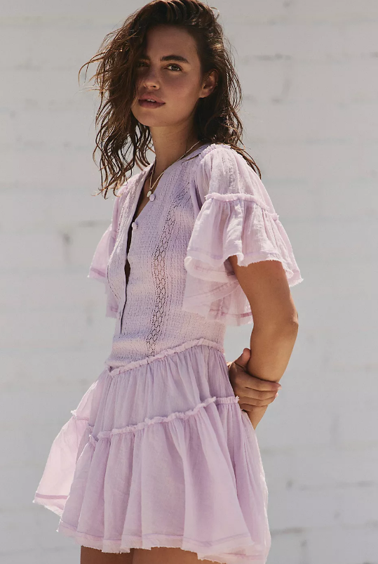 A woman with wavy brown hair stands against a light-colored wall, wearing a French Inspired Summer Dress in light purple with short sleeves, tiered ruffle details, and holding her left wrist with her right hand.
