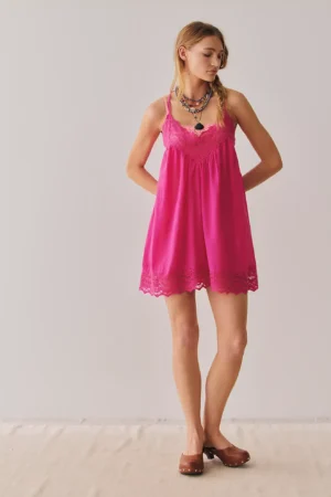 A woman stands against a plain light background, wearing the Intimately Starstruck Mini Slip—a bright pink lace-trimmed dress—paired with brown platform clogs. She has long braided hair and finishes the look with layered necklaces.