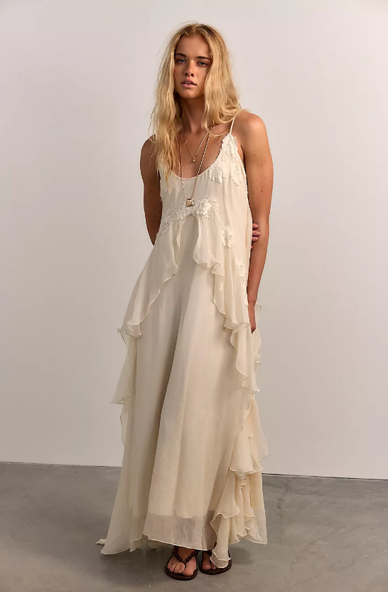 A woman with long, wavy blonde hair stands against a light-colored wall, wearing a sleeveless, floor-length off-white dress with ruffled layers and simple sandals—neutral shades defining summer style.