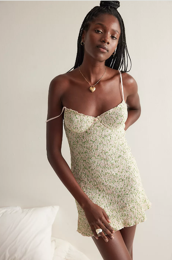 A woman stands indoors wearing a light-colored, floral mini slip dress with thin straps. She has long braided hair, accessorized with a necklace and ring, embodying neutral shades defining summer as she poses against a plain background near pillows.