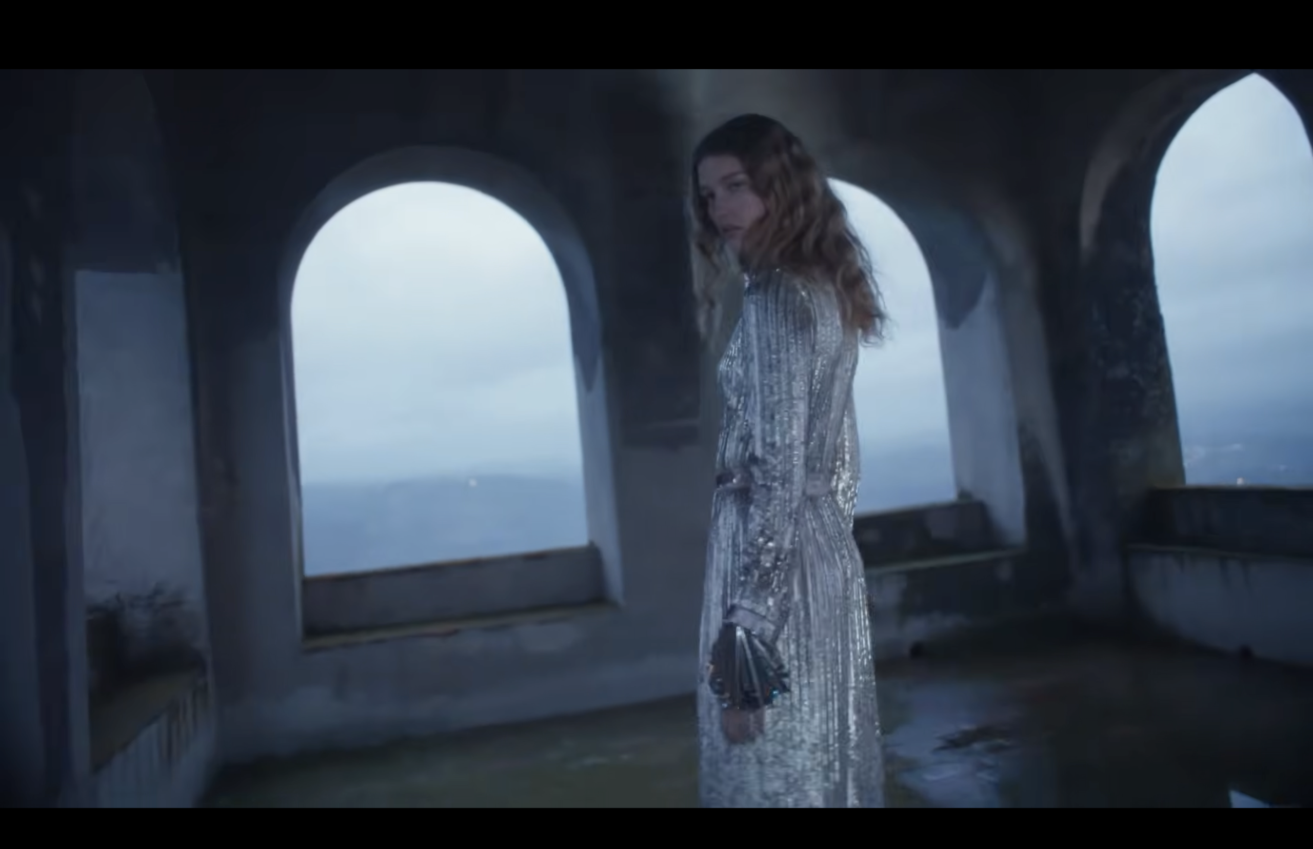 A person in a long, silver, reflective dress stands inside a large, stone structure with arched windows. The sky outside is overcast, and the floor appears wet.