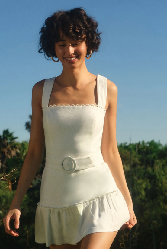 A person with short curly hair, wearing a sleeveless white vacation dress with a belt and ruffled hem, stands outdoors in sunlight, surrounded by greenery and blue sky—capturing the effortless charm of Style Insiders on the Amalfi Coast.