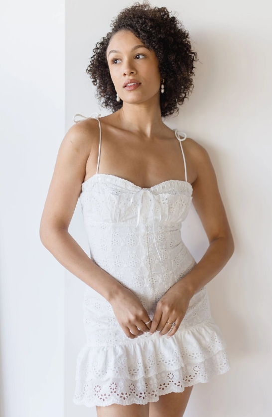 A woman with curly hair wears a white, sleeveless, eyelet dress—perfect for beach dresses—with thin straps and a ruffled hem. She stands indoors against a white wall, looking slightly to the side.