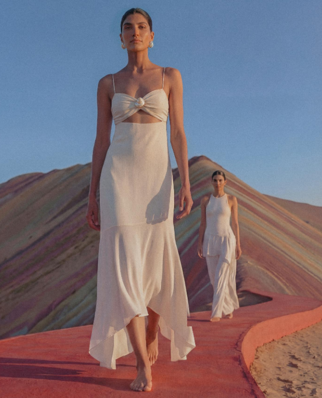 Two women in white vacation dresses walk barefoot on a red path before colorful, striped hills under a clear blue sky. The focus is on the woman in the foreground, with the second woman further behind.