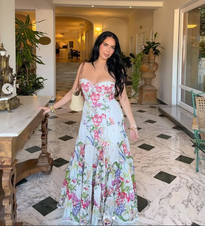 A woman with long dark hair wearing a white floral summer dress stands indoors on a marble floor, one hand resting on a table. The setting features plants, decorative items, and soft lighting—perfect for brunch or a day of shopping.