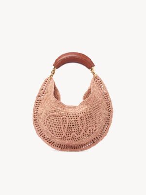 A blush pink crocheted handbag with a curved shape, brown leather handle, and "Chloé" stitched on the front, inspired by the Chloé Château Treasures Embellished Textured-leather Shoulder Bag, photographed against a plain white background.
