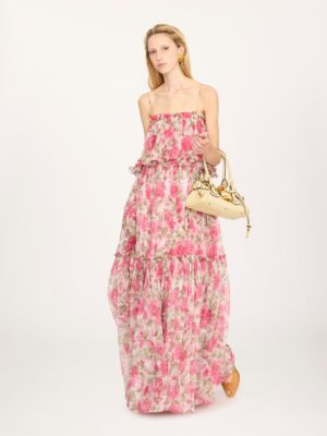 A person wears the Chloe Thin-Strap Long Dress in Silk Mousseline, featuring a long, pink floral design. They carry a beige handbag and wear brown shoes against a plain white background.