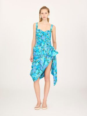 A person stands facing forward, wearing a sleeveless, blue floral dress with ruched detailing—reminiscent of the Chloe Long Sleeve Mini Dress in Lace—with a large side bow and an asymmetrical hemline. They have straight, shoulder-length hair and are wearing sandals.