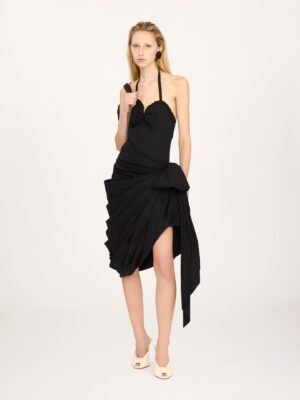A person with long blonde hair wears a Chloe Sleeveless Long Dress in Silk Charmeuse, featuring an asymmetrical halter-neck with ruched and draped details, paired with white open-toe heels, standing against a plain light background.