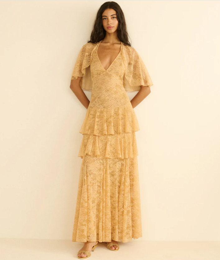 A woman stands against a plain beige wall wearing a floor-length, tiered, light yellow lace dress with short flutter sleeves—a perfect choice for those seeking dress styles that cover arms. She has long, wavy dark hair and is wearing sandals.