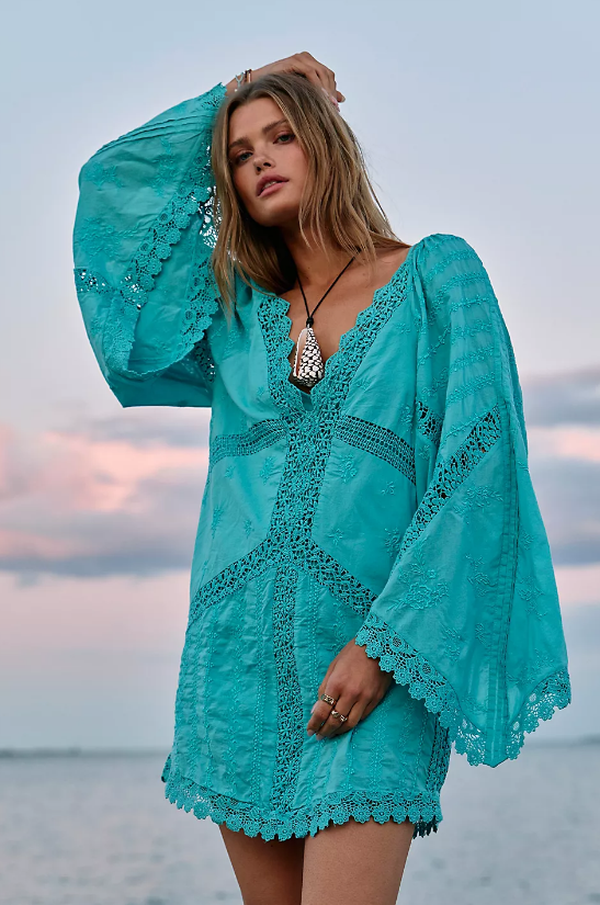A person stands outdoors near water, wearing a turquoise lace dress with wide, flared sleeves—a perfect example of beach summer dresses 2026—and a black necklace with a large pendant. The sky is partly cloudy during sunset.