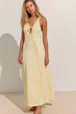 A woman stands barefoot against a plain background, wearing the Free-Est Full Of Sunshine cotton-linen mini dress with a front tie and lace detailing. She has long, wavy blonde hair and accessorizes with layered necklaces and bracelets.