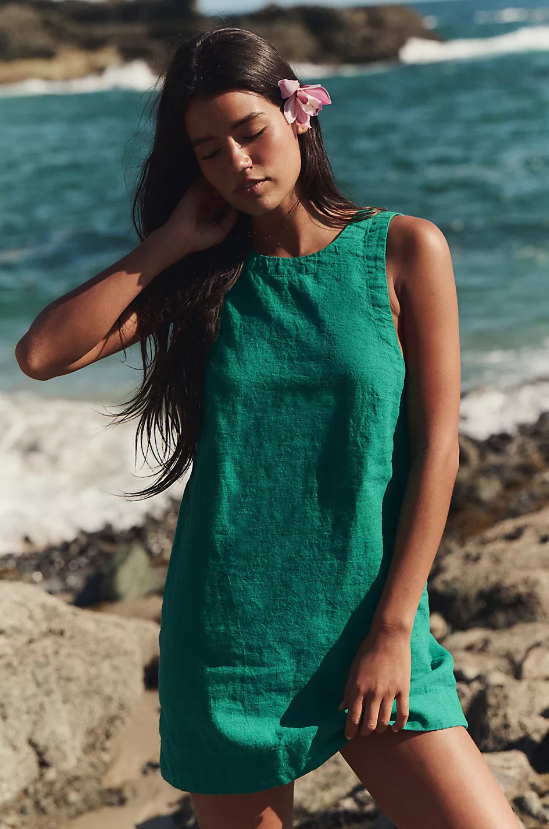 A woman models one of our beach summer dresses 2026 on a rocky shore, waves in the background. She wears a sleeveless green dress and a pink flower in her hair, eyes closed and one hand gently touching her neck.