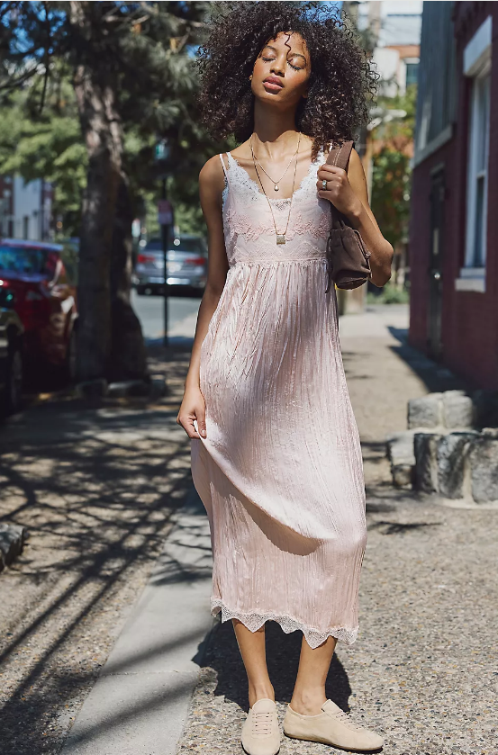 A person stands on a sunlit sidewalk wearing a light pink, sleeveless midi dress with lace details and tan shoes—a perfect example of styling flowy summer dresses. She holds a brown bag as trees, parked cars, and buildings fill the background.