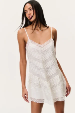 A woman stands against a plain light background, wearing a delicate white lace slip dress with thin spaghetti straps and sheer lace panels, reminiscent of the romantic style found in the LoveShackFancy Khloe Cotton Gingham Mini Dress.