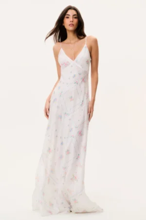 A woman wearing a long, white maxi dress with thin straps and a V-neckline featuring a pastel floral patchwork pattern, reminiscent of the romantic LoveShackFancy Madiya Crochet Cotton Mini Dress, stands against a plain light background.