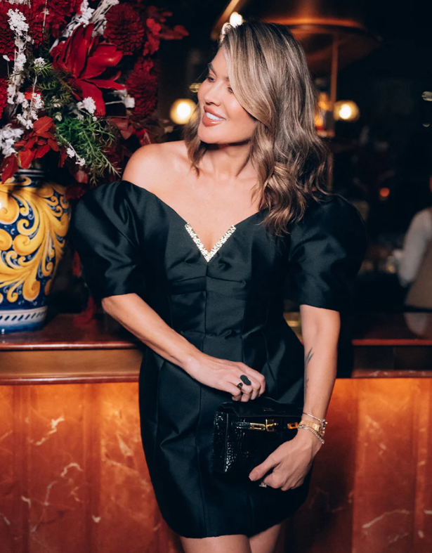 A woman with wavy, shoulder-length hair poses indoors, wearing an off-the-shoulder black dress featuring puffed sleeves—a chic example of dress styles that cover arms. She holds a black clutch and stands near a large vase of red flowers, bathed in warm lighting.