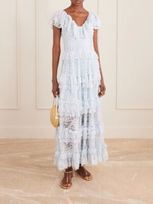 A person wearing the Sea Gracelyn Embellished Lace Maxi Dress in light blue, with ruffle details and short sleeves, stands indoors on a marble floor, holding a small round woven handbag and wearing flat metallic sandals.