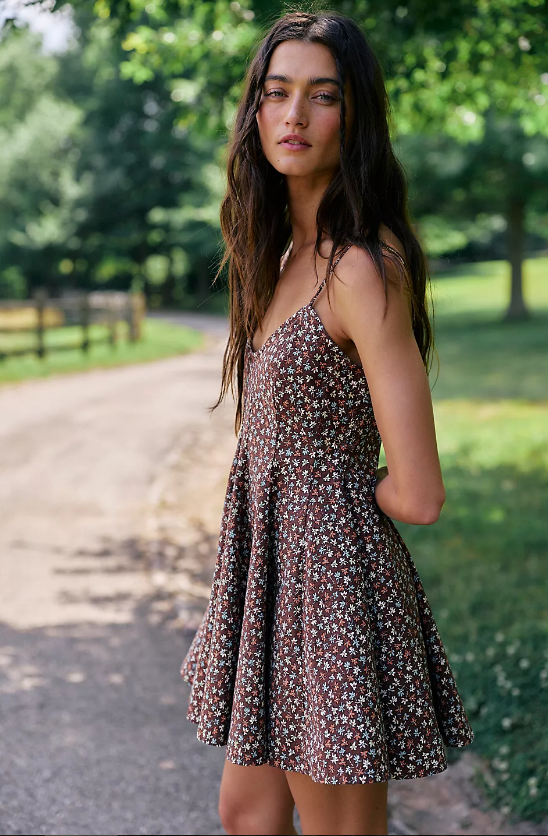 A woman with long brown hair stands on a path in a park, wearing a sleeveless, brown floral dress reminiscent of styles from the summer wishlist of any fashion editor. Trees and a wooden fence are visible in the background on a sunny day.