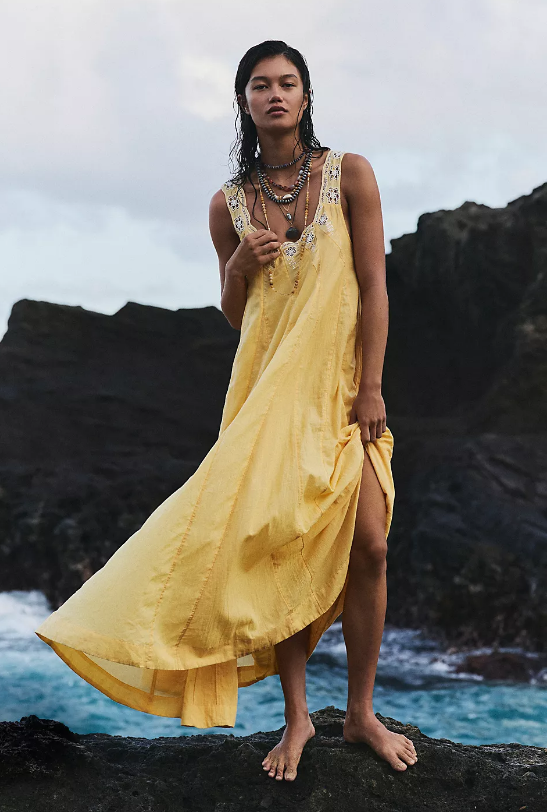 A person stands barefoot on rocks by the ocean, wearing a long, sleeveless yellow dress from Free People dresses with decorative trim at the neckline and several layered necklaces. The dress flows in the wind, capturing effortless summer fashion. The background includes water and rocky cliffs.