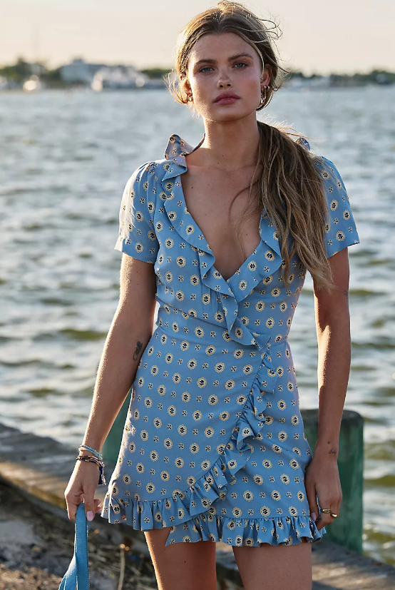 A woman stands outdoors near water, wearing a light blue, short-sleeved wrap dress with a floral pattern and ruffle details—a style that would top any fashion editor’s summer wishlist. Her hair is tied in a loose ponytail, and she is holding a small blue item in her left hand, perfectly capturing the effortless elegance of Free People dresses.