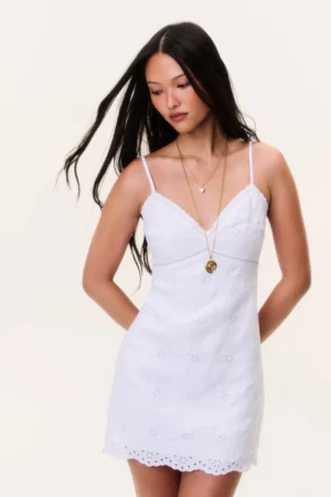 A woman with long dark hair wears the LoveShackFancy Becklyn Eyelet Linen Mini Dress, featuring thin straps and a scalloped hem. She accessorizes with two layered necklaces, standing against a plain light background and looking down to the side.