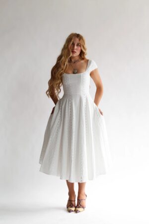 A woman with long wavy hair wears the Wild Rose & Sparrow The Sylph Gown—a white, short-sleeved, midi-length dress with eyelet details and a fitted bodice. She pairs it with purple sandals and layered necklaces against a plain white background.