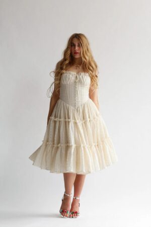 A woman with long, wavy blonde hair wears the Wild Rose & Sparrow The Pirouette Gown—a cream-colored, tiered dress with a fitted bodice and ruffled skirt. She stands against a plain white background, wearing open-toed heels.