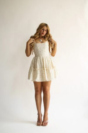 A person stands against a plain white background wearing the Wild Rose & Sparrow The Juliette Dress, a sleeveless, light-colored, tiered mini dress with small dark patterns and clear-strap sandals. They have long wavy hair and are holding two sections of it.